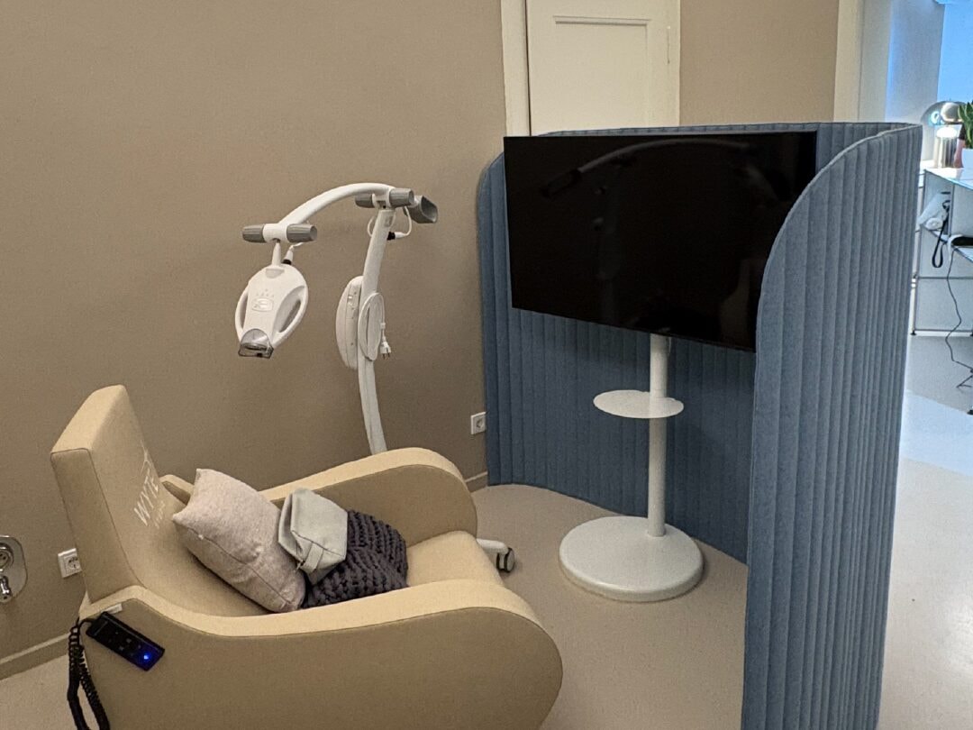 A WYTE Studio treatment room: a cream reclining chair with a cushion and a knitted blanket, the whitening lamp on a stand beside it, and a television behind a blue acoustic screen.